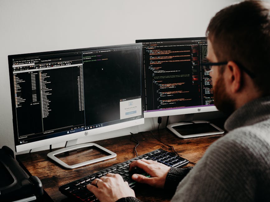 A person with dark hair, glasses, and a beard is seated at a wooden desk working on a computer. They are wearing a grey sweater and are using a keyboard with their right hand while their left hand res